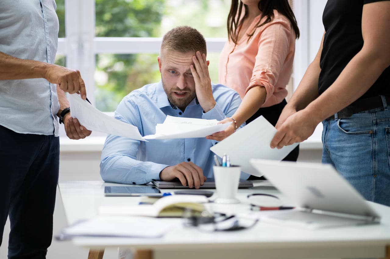 Stresset mand på kontor omgivet af kollegaer med papirer – behov for stressbehandling Lemvig.